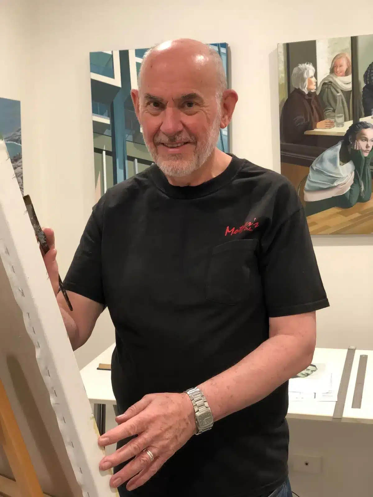 A man holds a paintbrush, standing in front of a colorful painting on an easel.