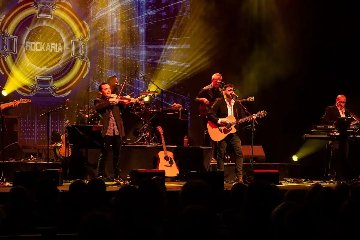A band performs on stage with a large screen displaying visuals behind them.