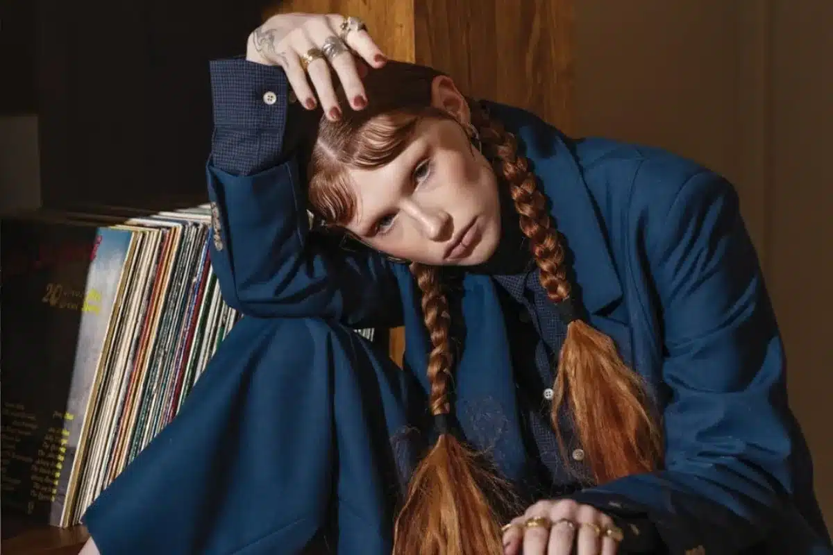 A woman with red hair sits on a chair beside a record player, enjoying music in a cozy setting.