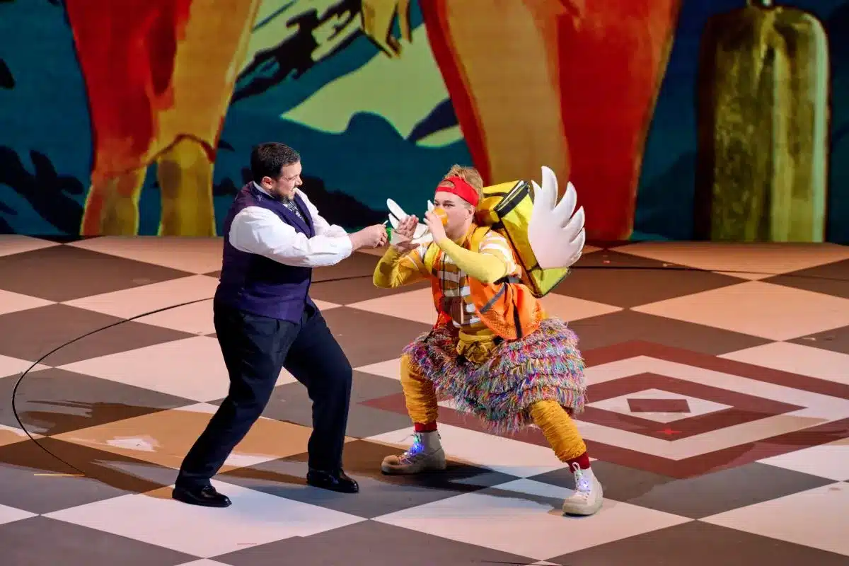 Two men in colorful costumes perform on stage, accompanied by a bird perched nearby.
