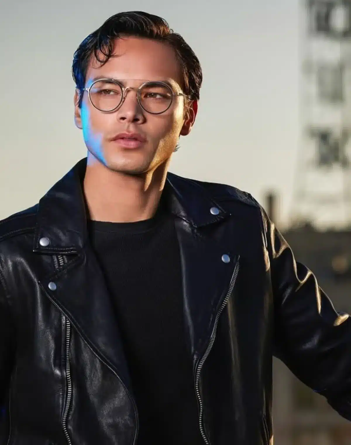 A man with glasses wearing a black leather jacket, standing confidently against a neutral background.