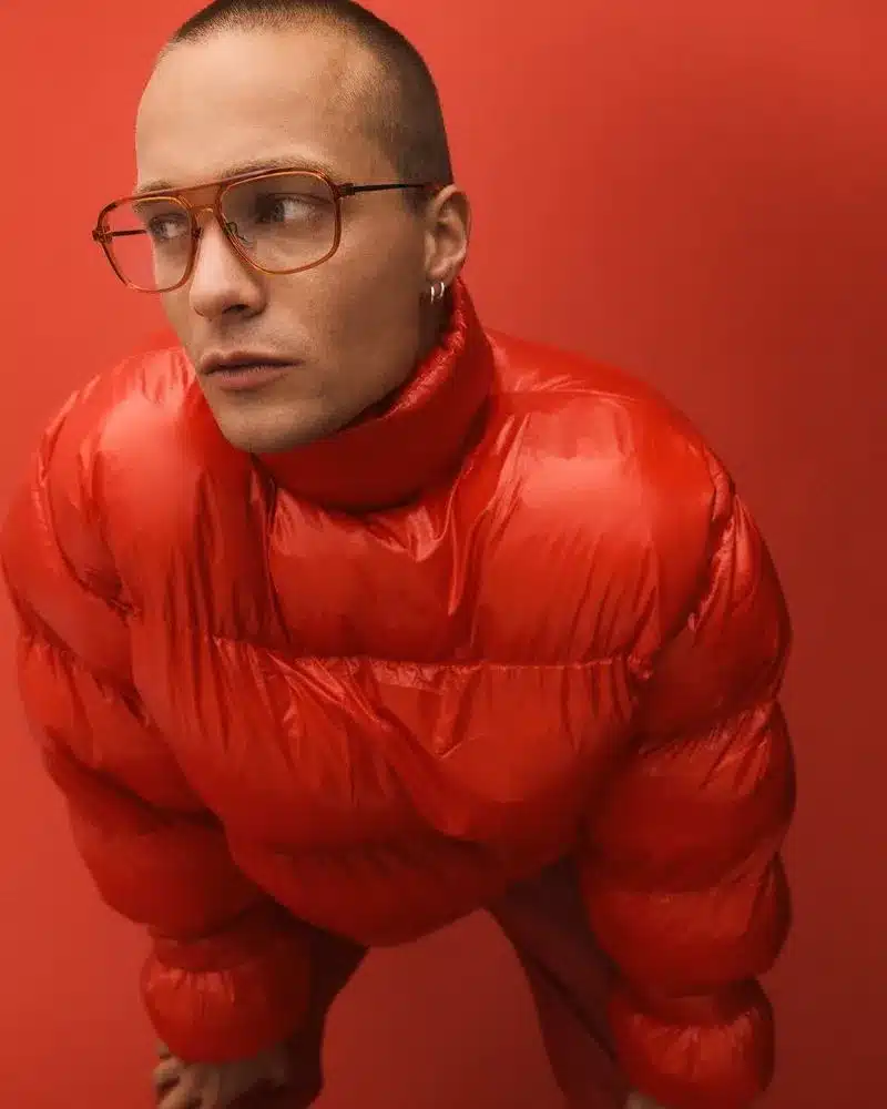A man with glasses wearing a bright red puffy jacket stands against a neutral background.