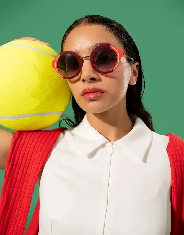 A woman wearing sunglasses holds a tennis ball, ready for a game on a sunny day.