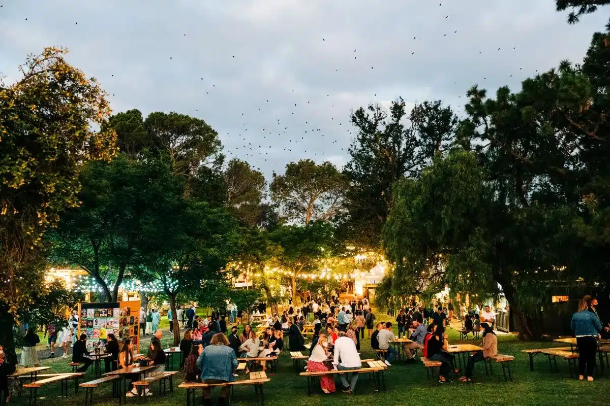 A diverse crowd of people seated at tables enjoying a sunny day in a park.