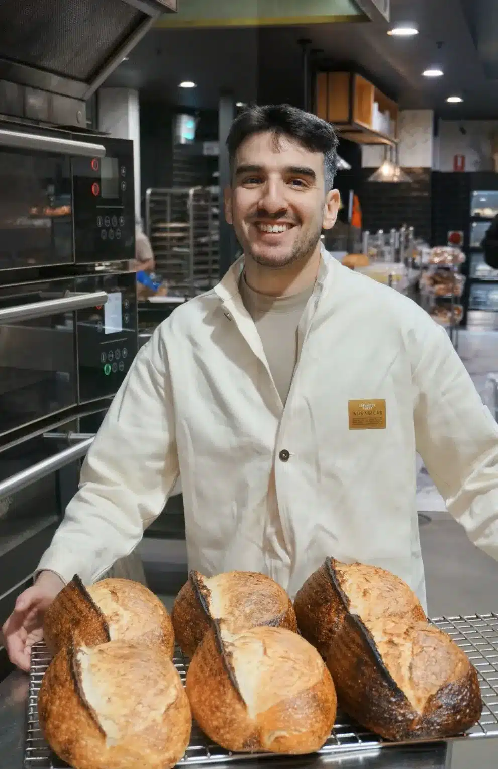 A man in a white coat holds a tray filled with freshly baked bread, smiling as he presents it.