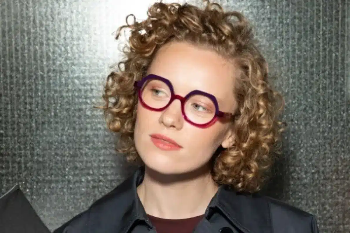 A woman with curly hair and glasses smiles at the camera, showcasing her stylish look.
