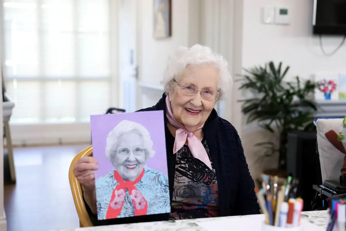 An elderly woman proudly holds a painting of herself, showcasing her likeness and artistic expression.
