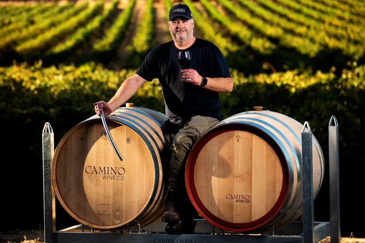 A man casually seated atop two stacked wine barrels in a rustic setting.