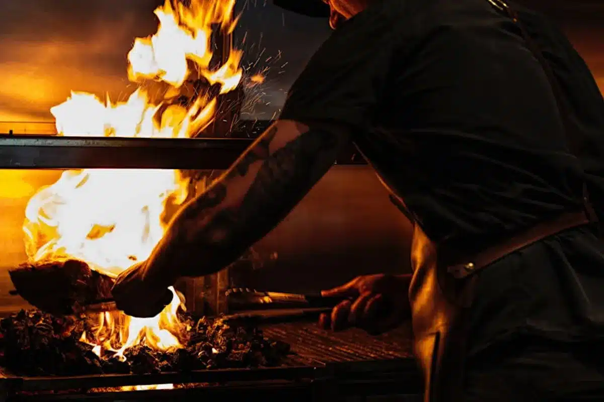 A man wearing a hat grills a steak on an outdoor barbecue, focusing intently on the cooking process.