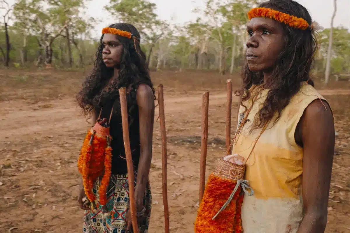 Two women wearing vibrant orange headdresses stand on a dirt surface, showcasing their cultural attire and surroundings.