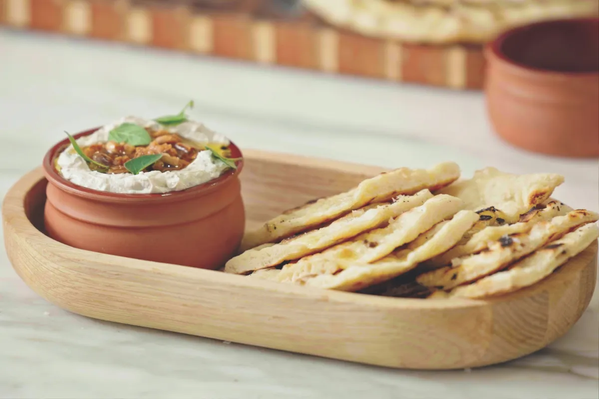 A wooden tray holds an assortment of bread alongside a small bowl of dip.