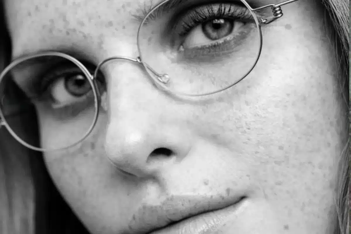 A woman with glasses and freckles gazes directly at the camera, smiling softly.