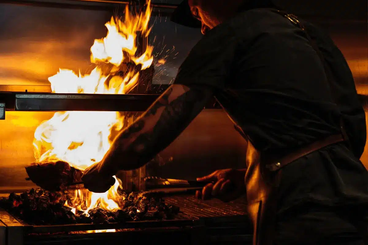 A man stands in a kitchen, watching a fire burning on the stove, with pots and utensils nearby.