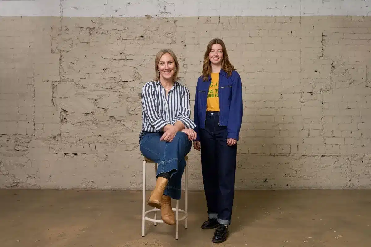 Two women seated on stools, smiling, in front of a textured brick wall.