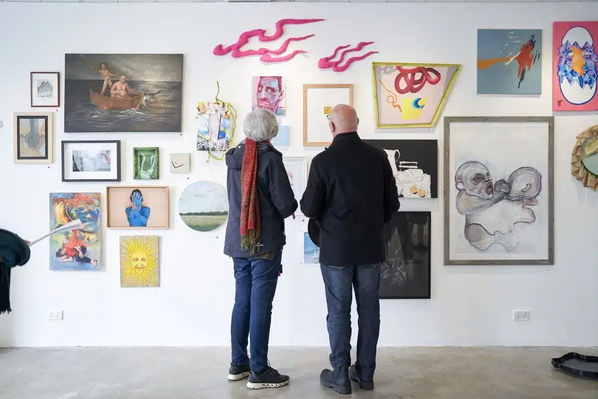 Two individuals observing artwork displayed on a wall in a gallery setting.