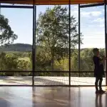 A person gazes out at a scenic landscape through large windows, standing in a well-lit interior space.