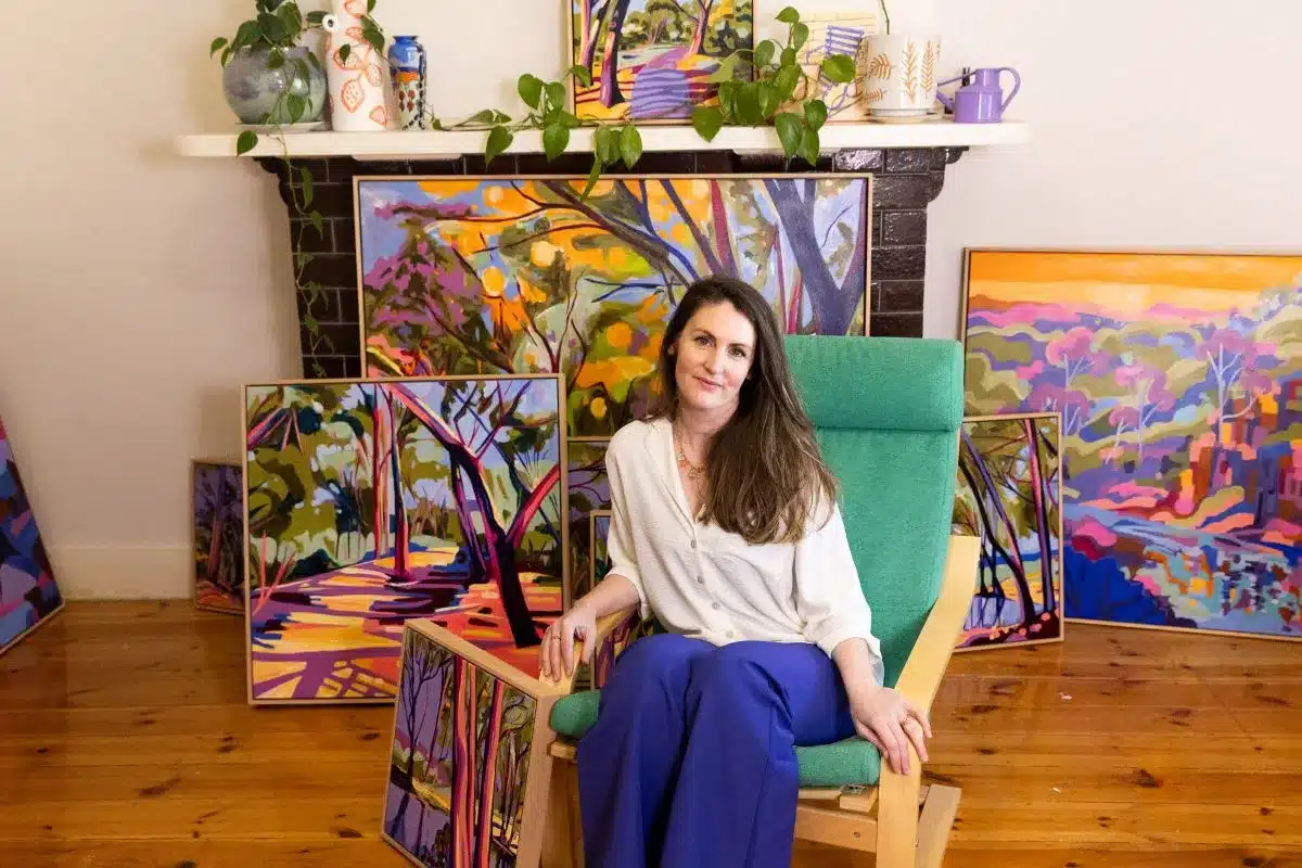 Artist sits on a green chair in front of her artwork.