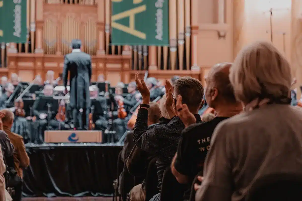 A large crowd gathers in an auditorium, attentively watching a conductor lead an orchestra performance.