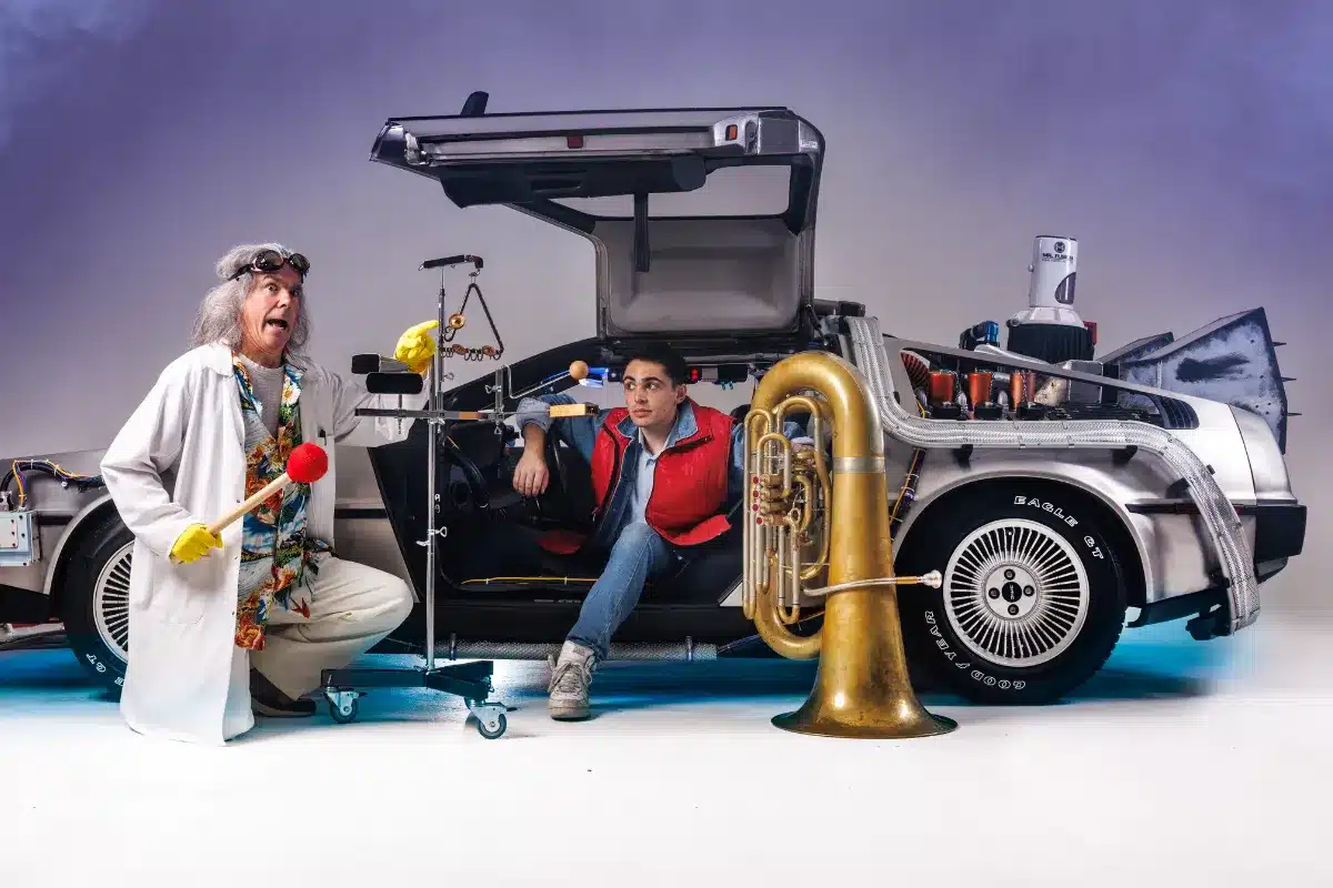 Two people stand in front of a car, one holding a trumpet, smiling and enjoying a sunny day.