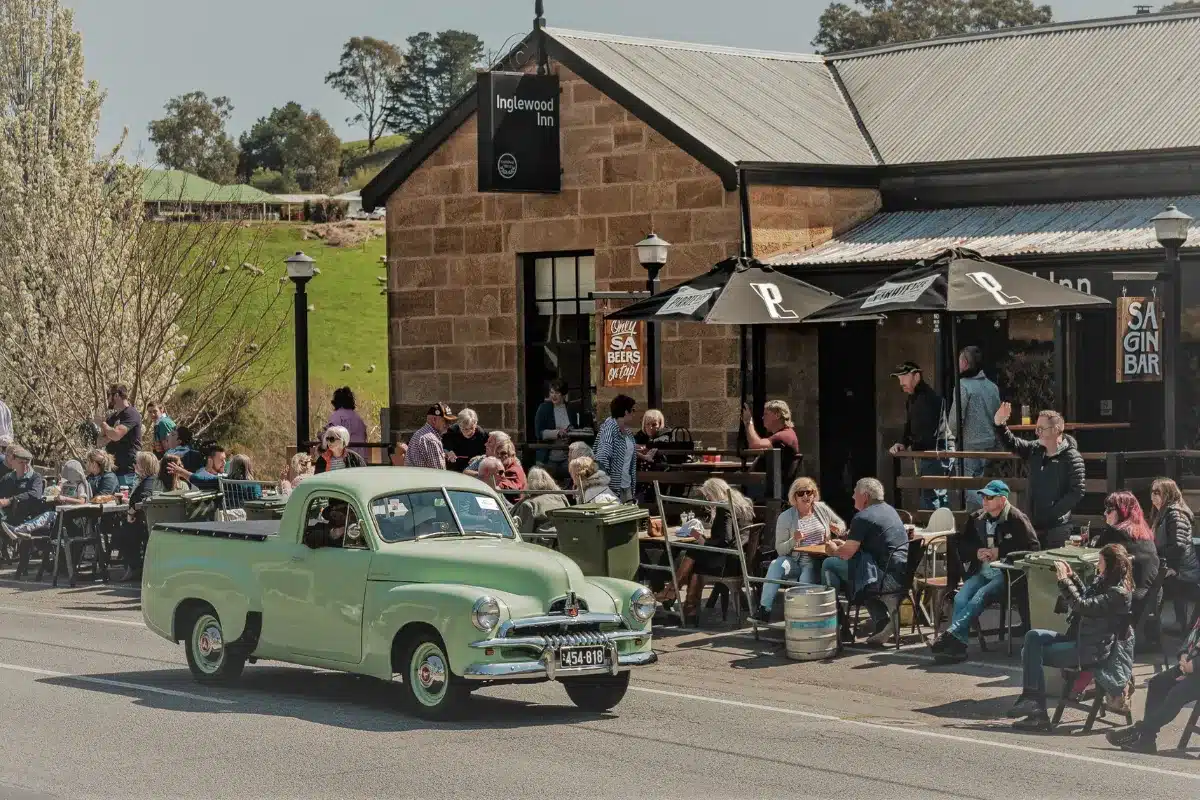 A green car drives down a street where people are seated outside, enjoying the day.