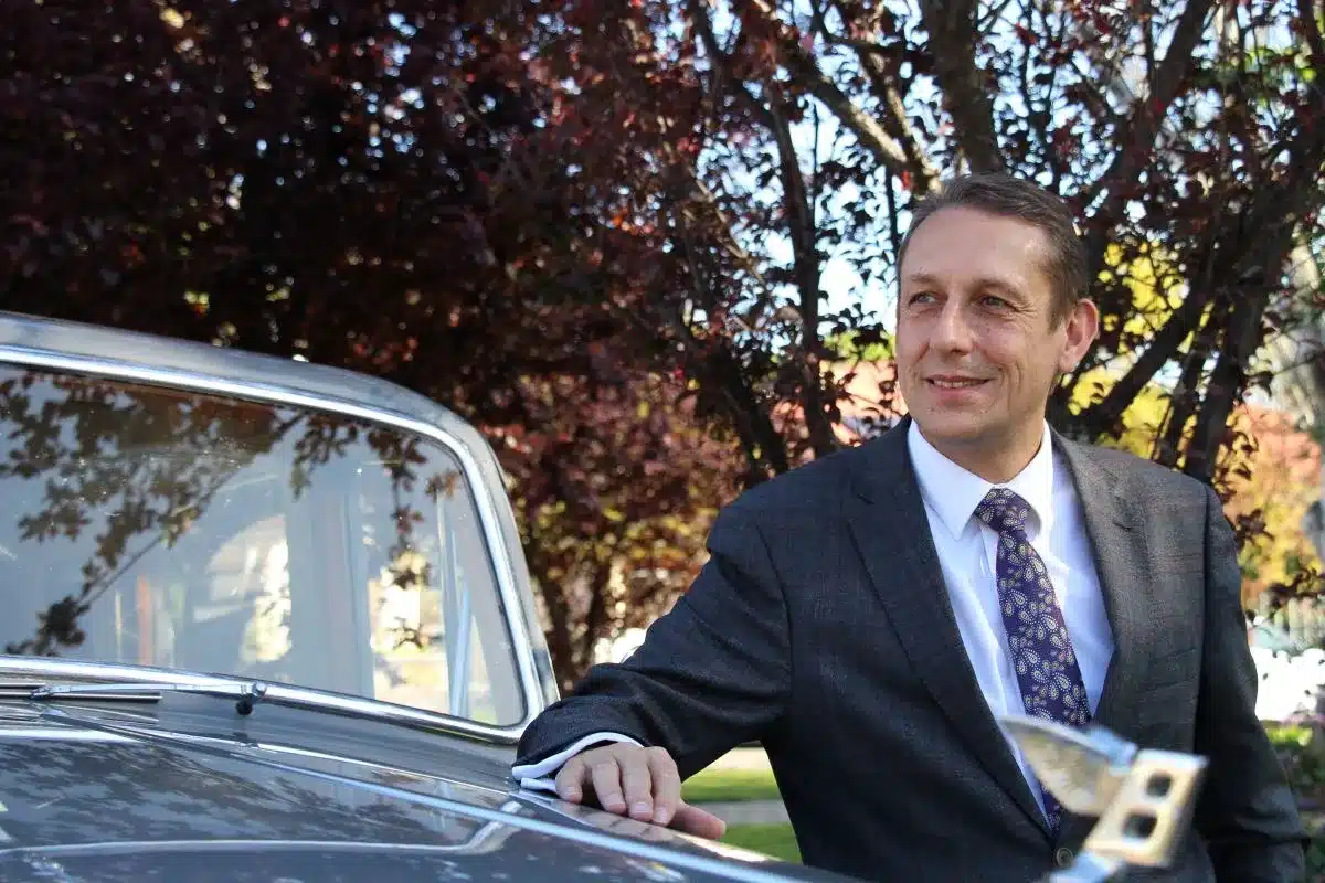 A man in a suit and tie leans against a classic car, exuding confidence and style.