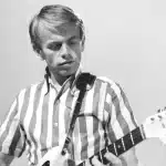 A man in a striped shirt passionately playing an electric guitar on stage.