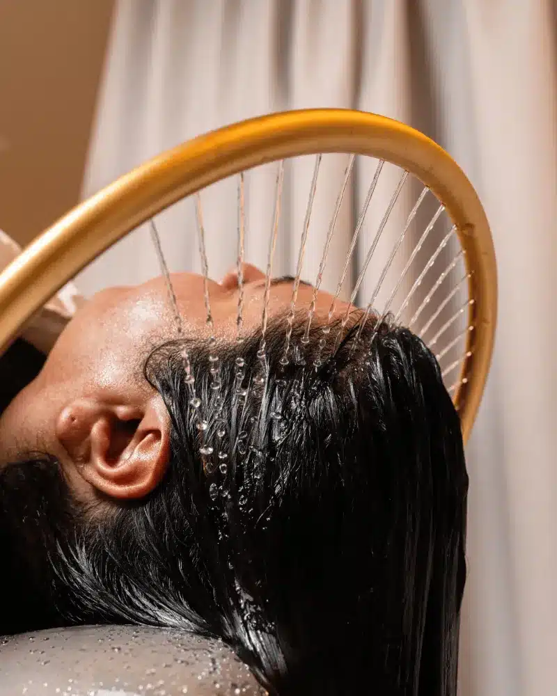 A woman relaxes as her hair is being washed at a salon, with water cascading over her head.