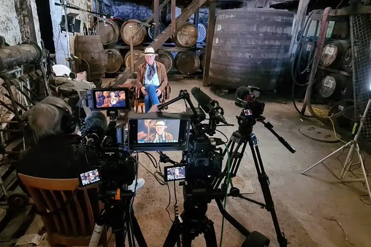 A man filming in a room, holding a camera while another camera is set up on a tripod nearby.