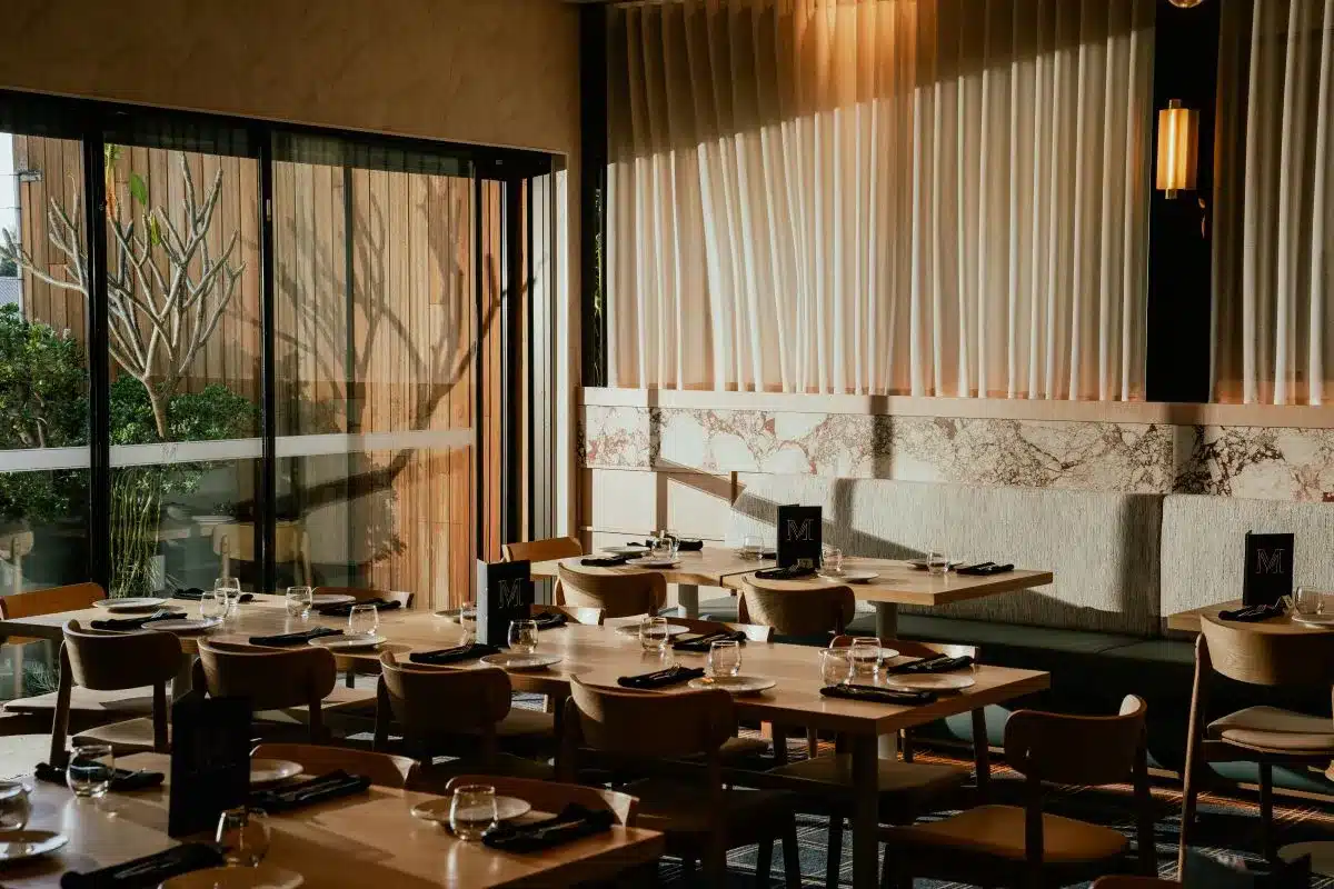 A restaurant interior featuring tables and chairs arranged near large windows, allowing natural light to fill the space.