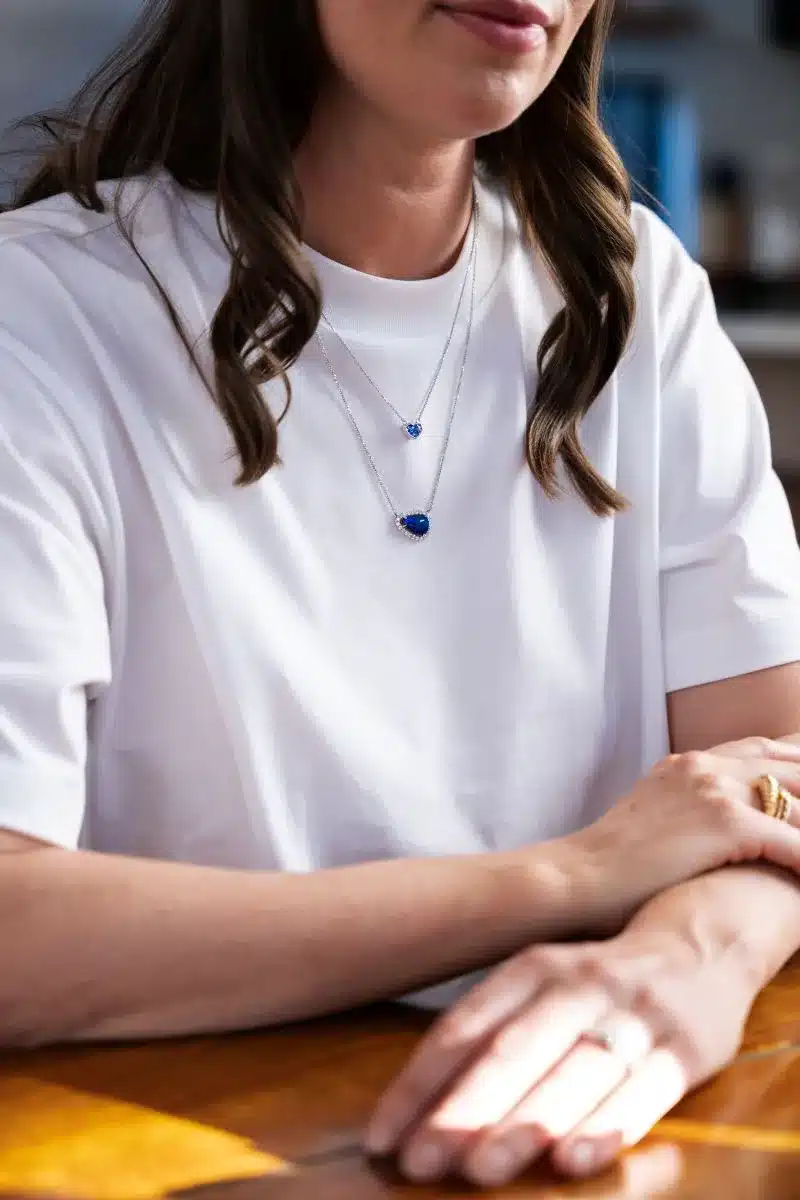 A woman in a white t-shirt wearing a blue necklace, smiling and looking towards the camera.