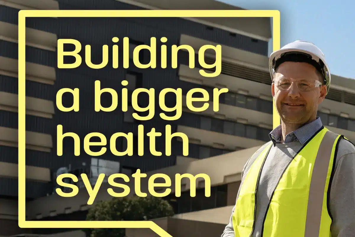 A man in a safety vest stands before a building displaying the phrase "Building a Bigger Health System."