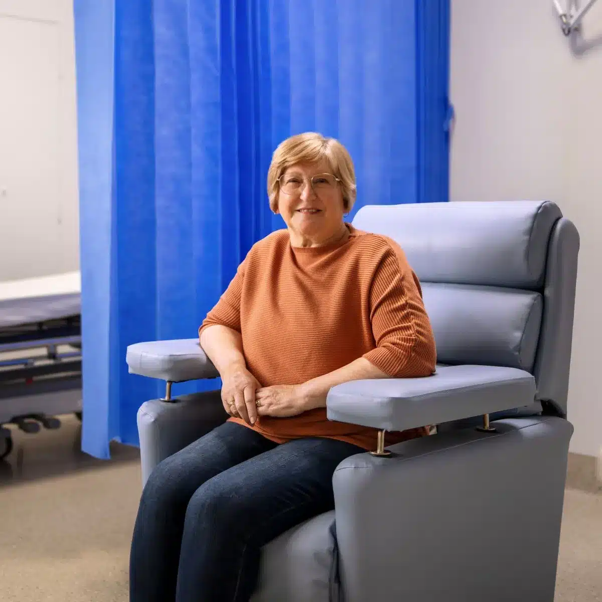 A woman sitting in a chair in a hospital room, looking contemplative and surrounded by medical equipment.