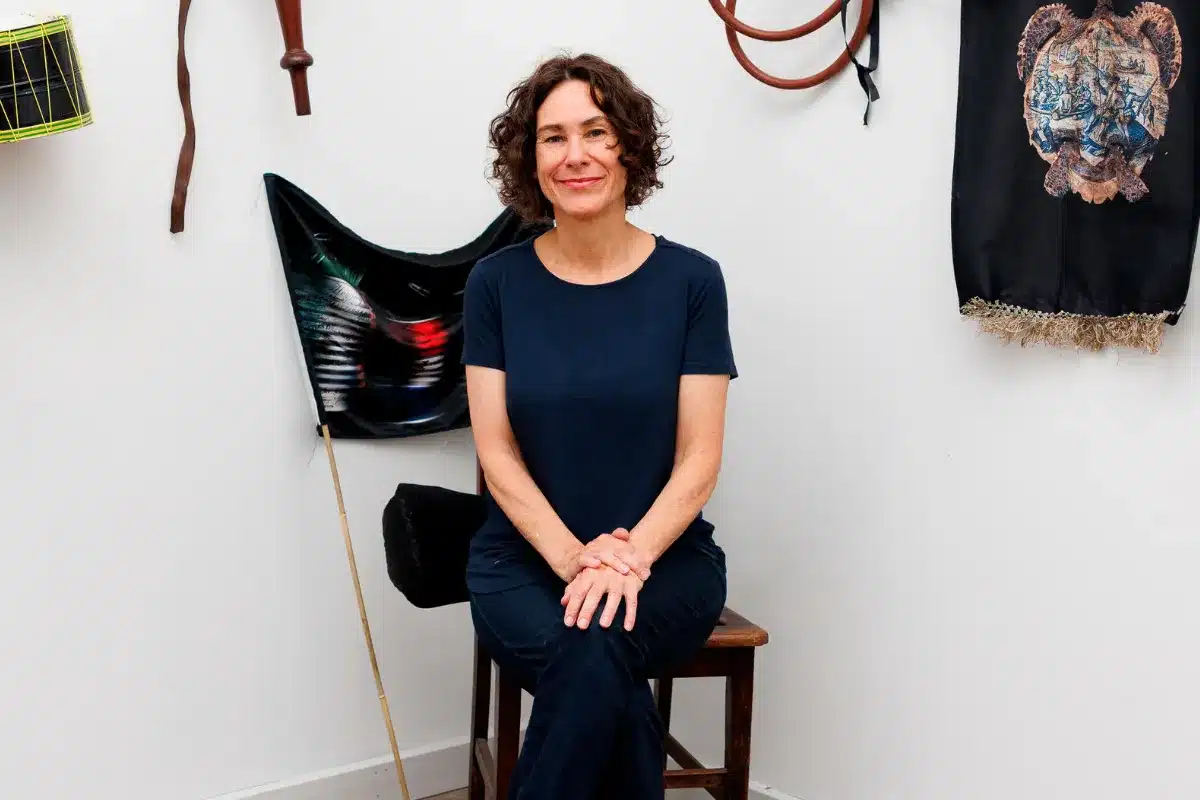 A woman seated on a chair, surrounded by various items displayed on a wall behind her.