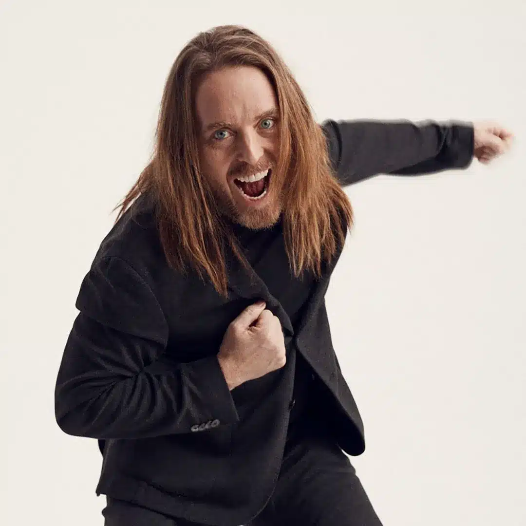 A man in a suit with long hair is joyfully jumping against a plain background.