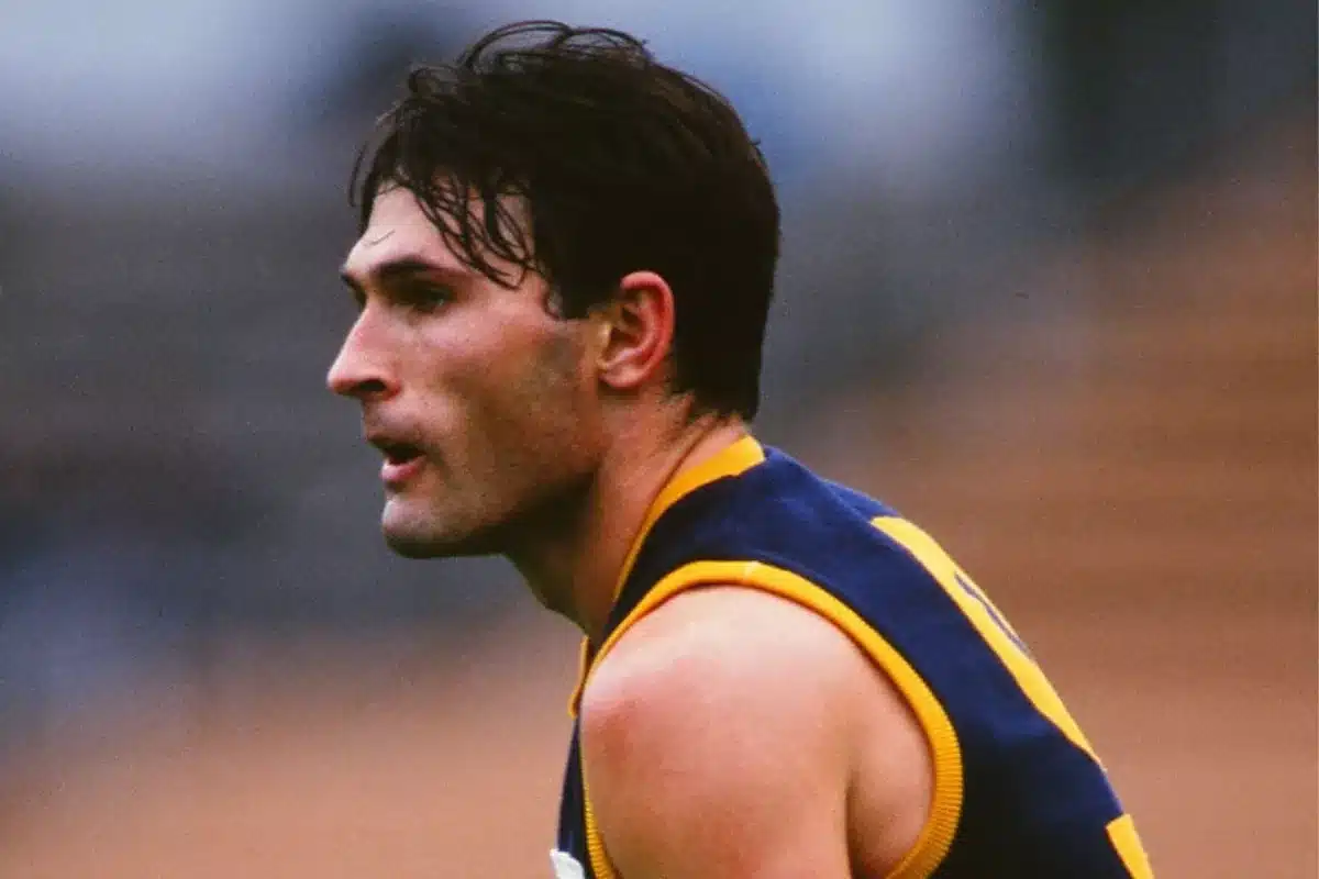 A male AFL player wearing a blue and yellow uniform stands on the field, ready for action.