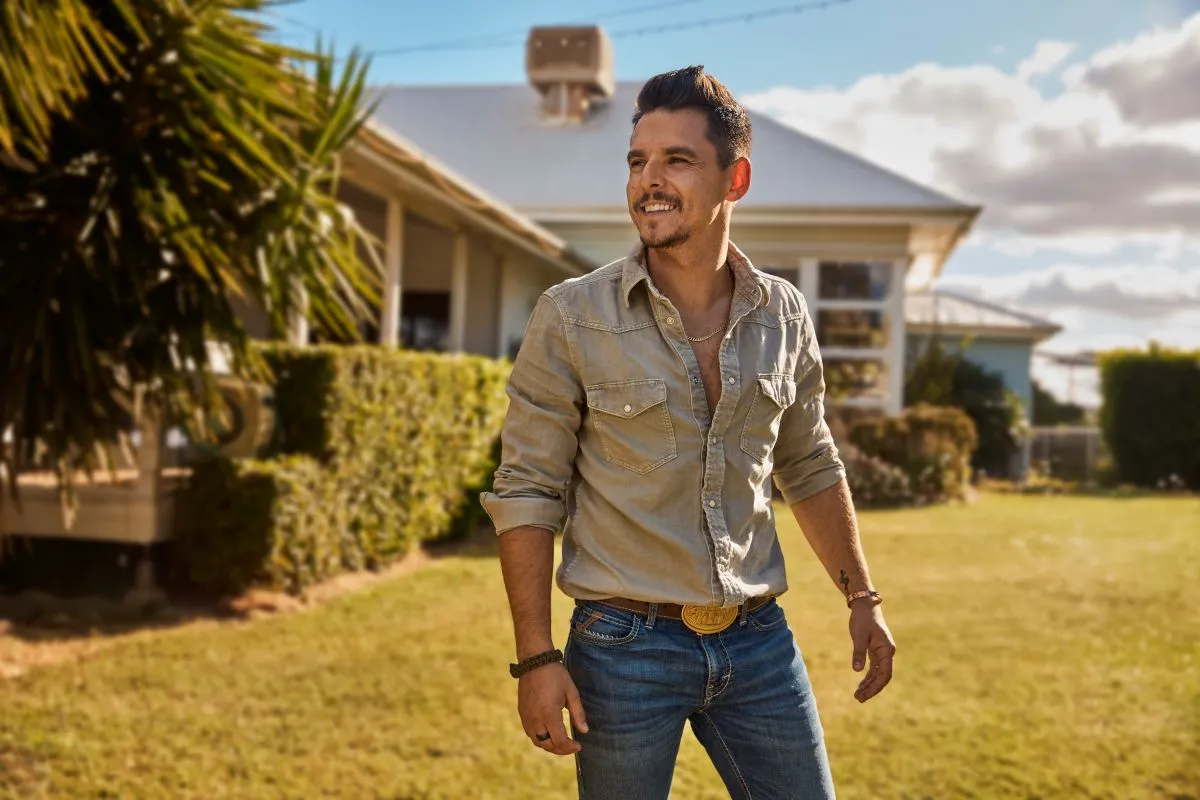 A man wearing jeans and a shirt stands in front of a house, smiling and looking towards the camera.
