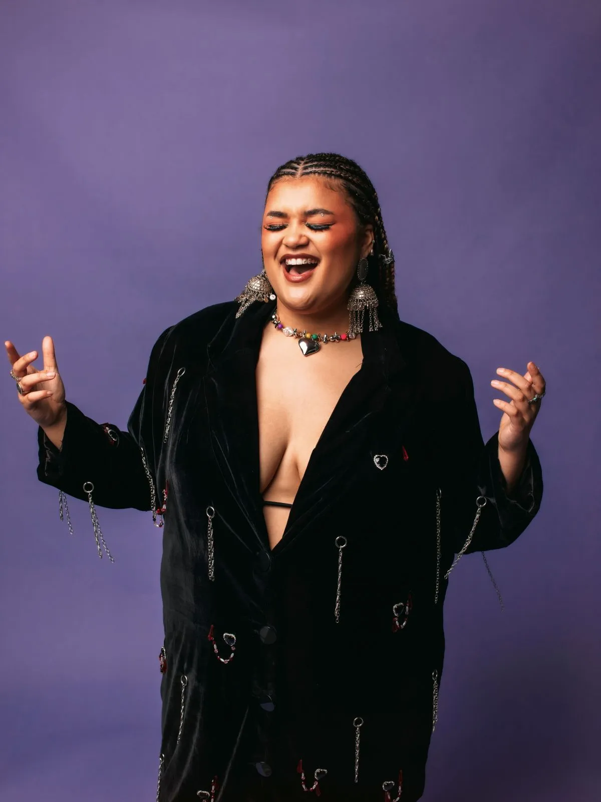 A woman wearing a black velvet jacket over a bra, posing confidently against a neutral background.