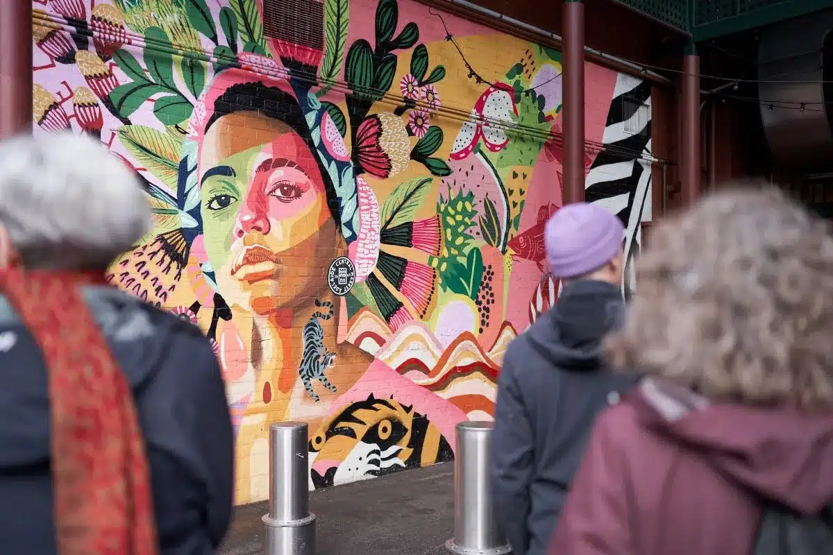 People stroll past a vibrant mural painted on a building, showcasing a blend of colors and artistic designs.
