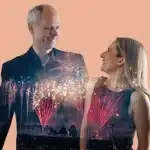 A man and woman stand together, smiling, as colorful fireworks illuminate the night sky behind them.