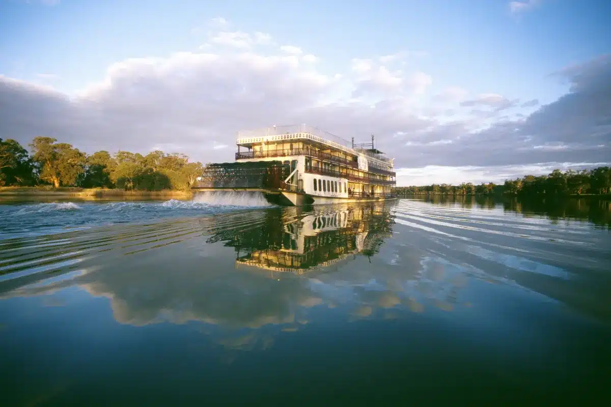 A river boat glides down a river, surrounded by lush trees in the background.