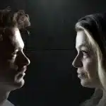 A man and woman gaze at each other in a dimly lit room, creating an intimate atmosphere.