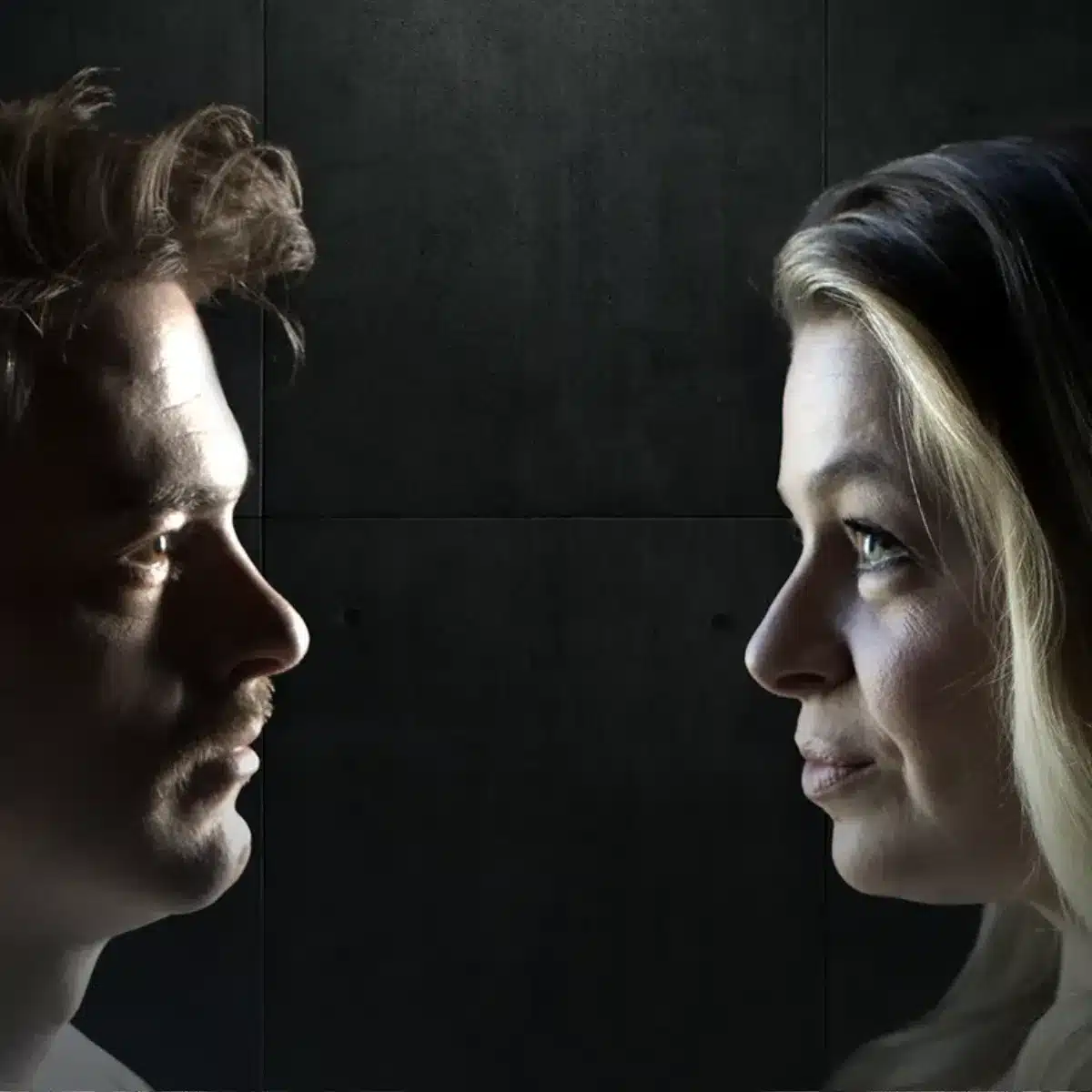 A man and woman gaze at each other in a dimly lit room, creating an intimate atmosphere.