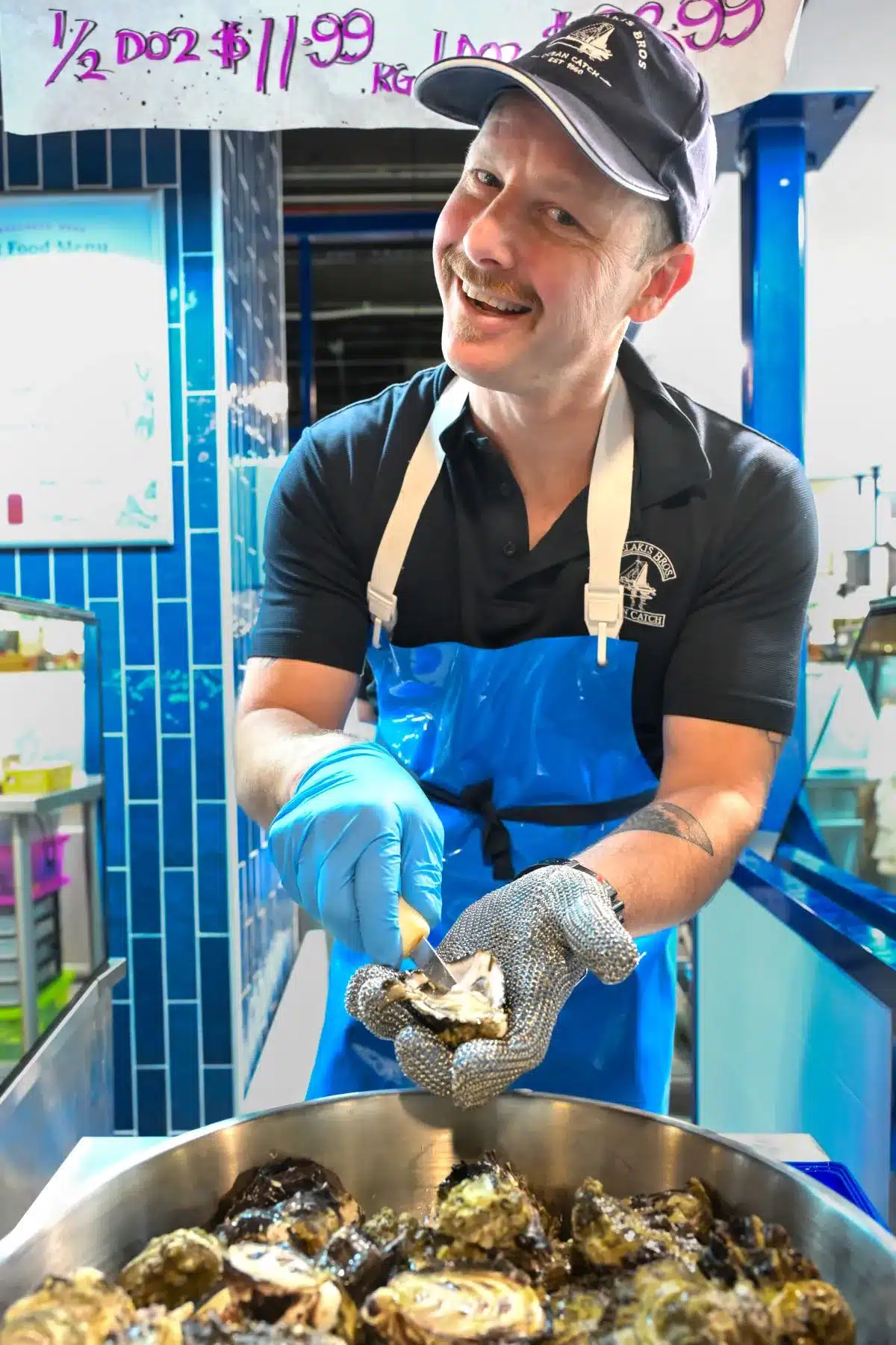 A man in a blue apron holds a bowl filled with fresh oysters, ready for serving or preparation.
