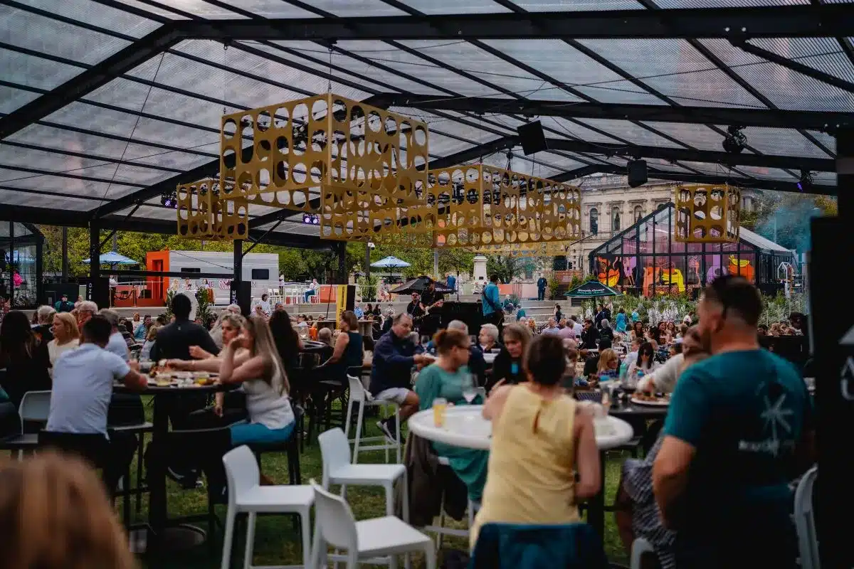 A large outdoor event featuring numerous people seated at tables, enjoying food and conversation under a clear sky.