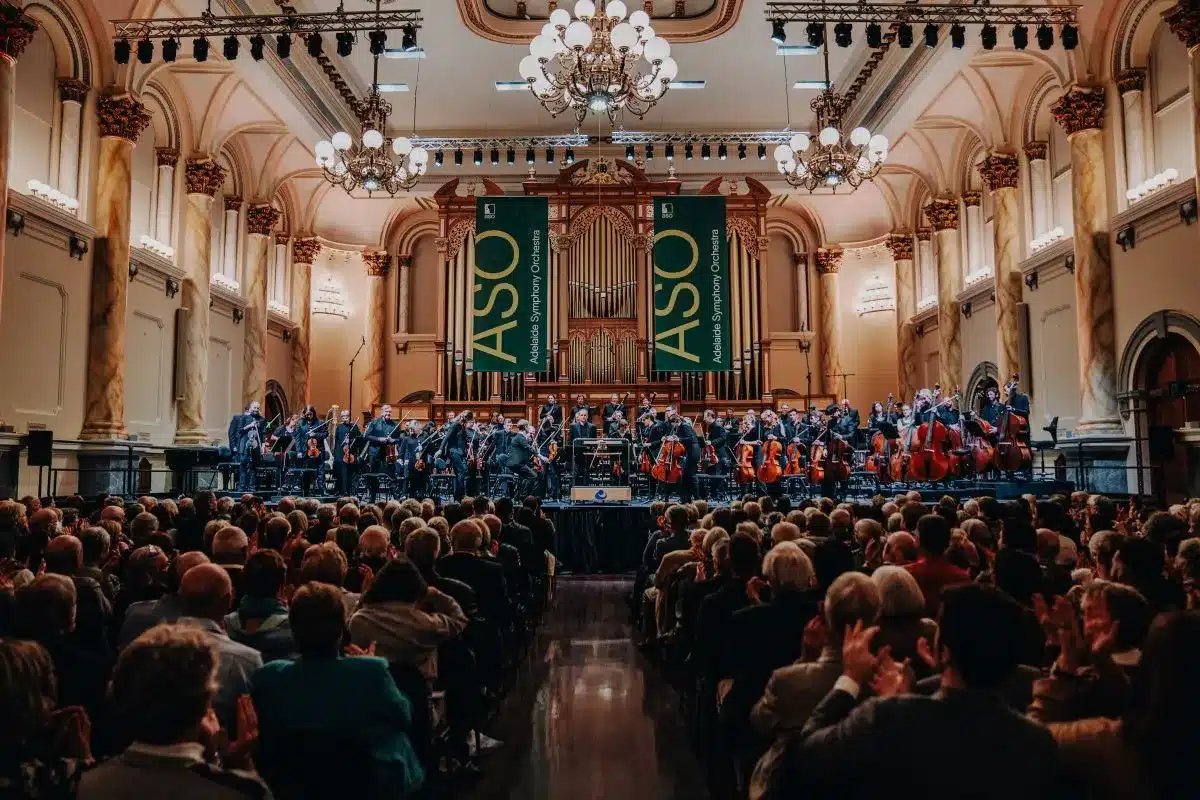 A large audience seated in a spacious hall, attentively watching a live orchestra performance on stage.