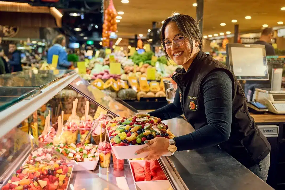 A woman holds a tray of colorful fruit while standing in a bustling market.