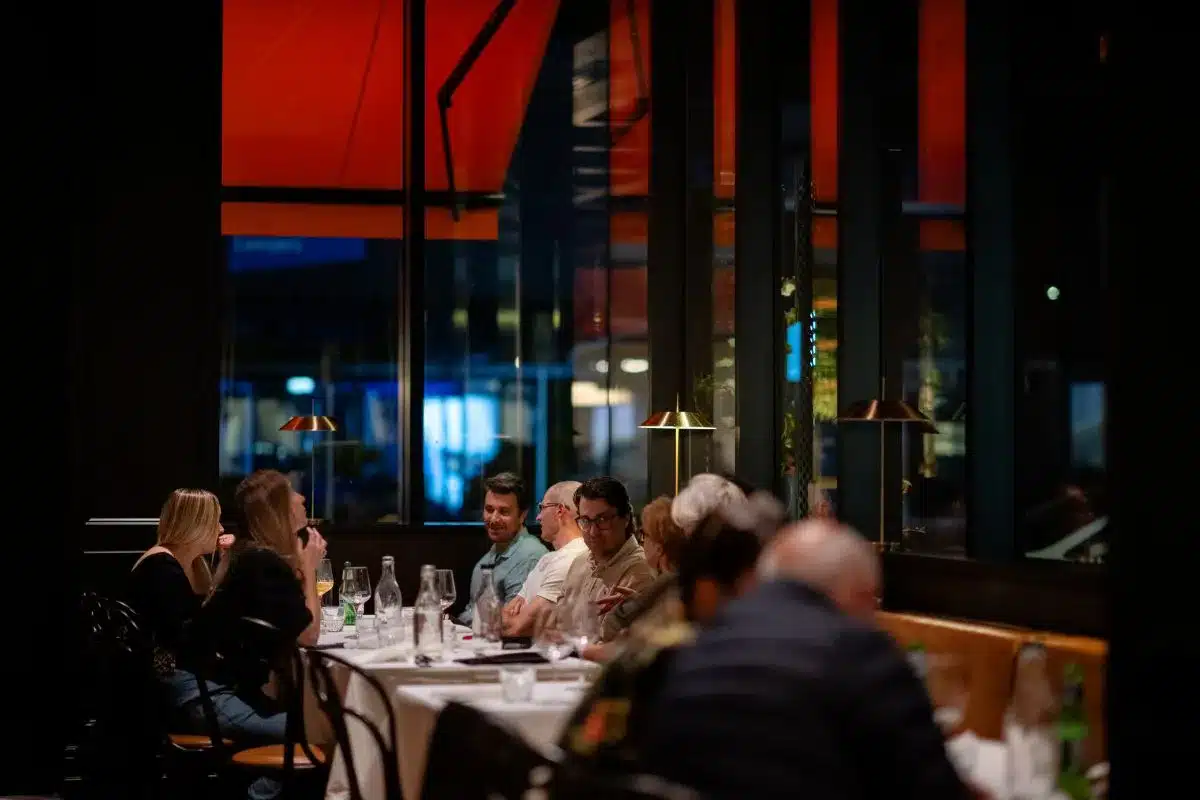 Diners seated at tables in a restaurant, enjoying meals by large windows that let in natural light.