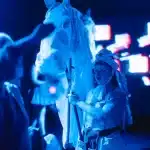 A person in a white costume stands beside a horse, gently holding its reins in a serene outdoor setting.