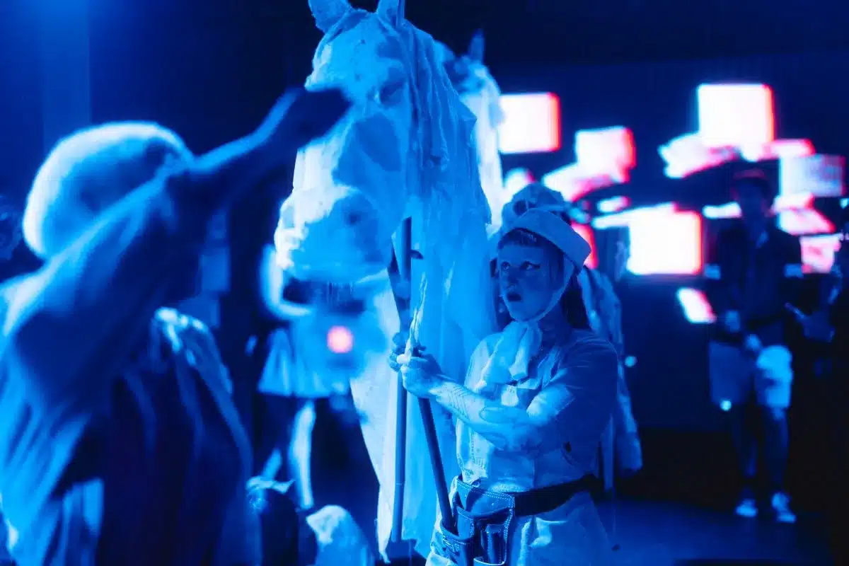 A person in a white costume stands beside a horse, gently holding its reins in a serene outdoor setting.