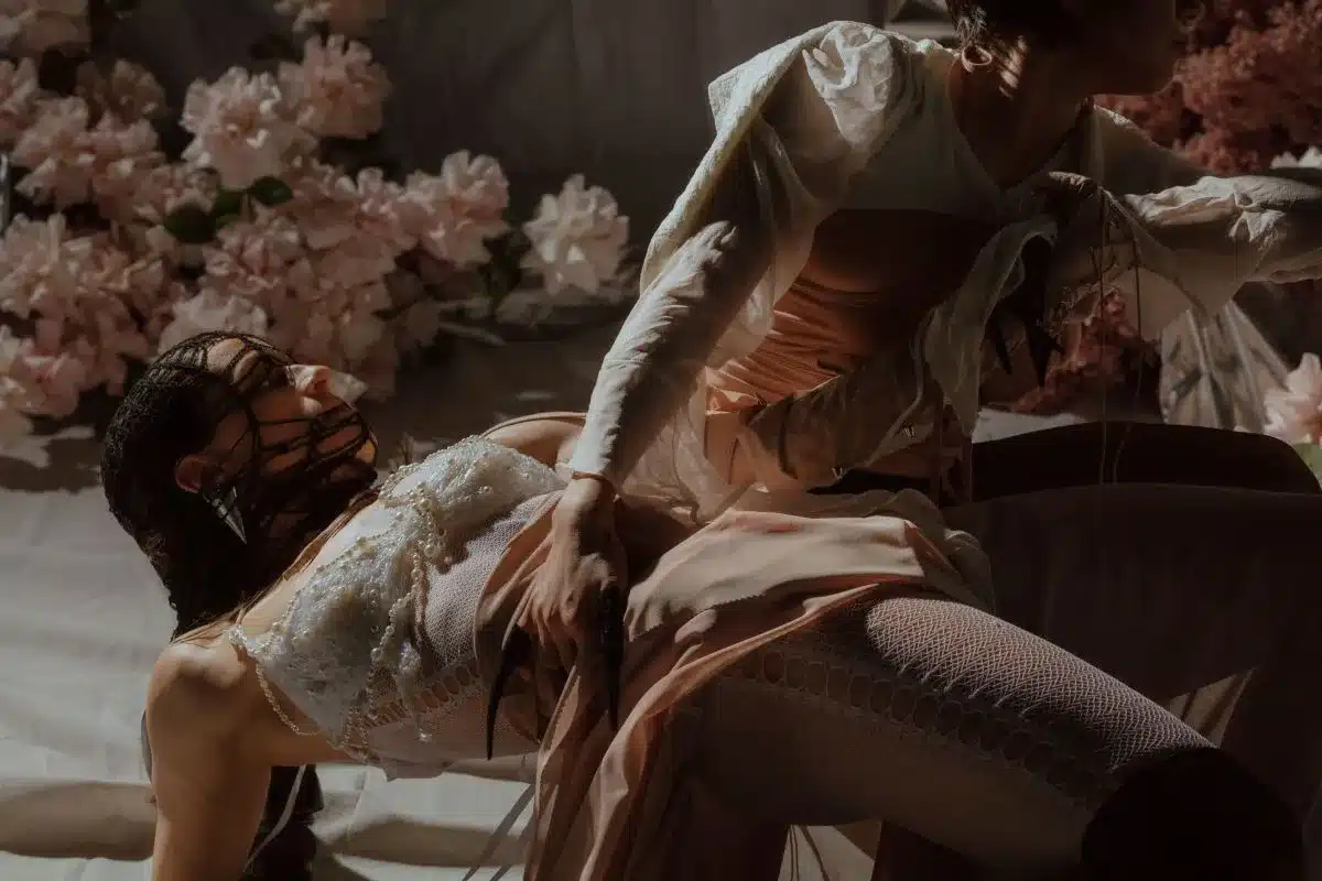 Two women in white dresses are lying on the ground, surrounded by grass and flowers, enjoying a peaceful moment together.